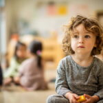 Four-year-old child sitting thoughtfully in a preschool classroom, holding a toy, representing early autism traits and developmental milestones.