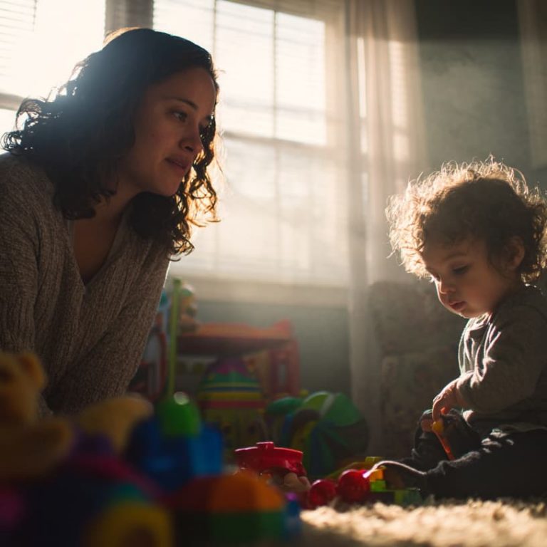 Mother gently observing her toddler playing with toys in a sunlit living room, representing early signs of autism awareness.